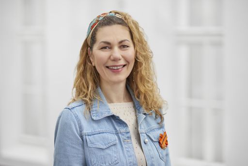 Lächelnde Frau mit lockigem blondem Haar, buntem Haarreif, Jeansjacke und orangefarbener Brosche vor hellem Hintergrund.