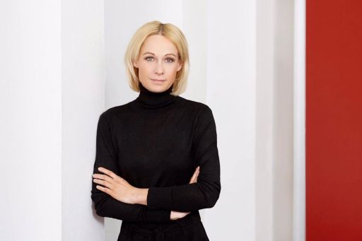 Blonde Frau in schwarzem Rollkragenpullover, Arme verschränkt, lehnt an weißer Wand, roter Hintergrund rechts.