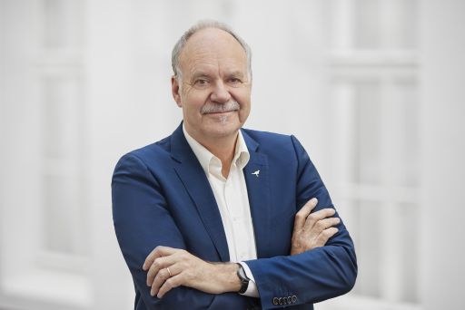 Älterer Mann mit grauem Haar und Schnurrbart, blaues Sakko, verschränkte Arme, vor hellem Hintergrund.