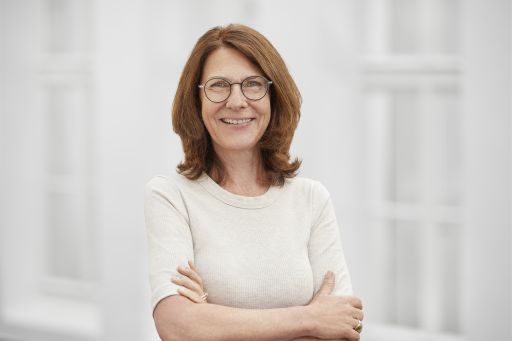 Frau mit roten Haaren, Brille und weißem Top, Arme verschränkt, lächelt vor hellem Hintergrund.