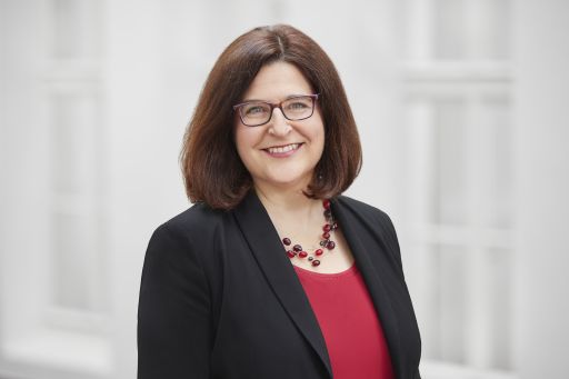 Frau mit braunem Haar und Brille, rotem Top, schwarzem Blazer und roter Perlenkette lächelt in die Kamera.