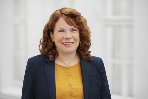 Frau mit roten Locken, gelbem Oberteil und blauem Blazer lächelt vor hellem Hintergrund.