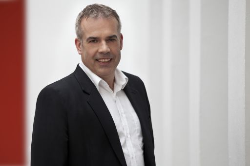 Mann mittleren Alters mit grauem Haar, schwarzem Sakko und weißem Hemd lächelt vor weißer Wand.
