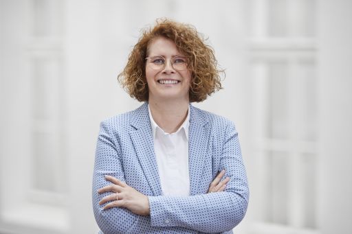 Frau mit roten Locken und Brille, in hellblauem Blazer, lächelt mit verschränkten Armen vor hellem Hintergrund.