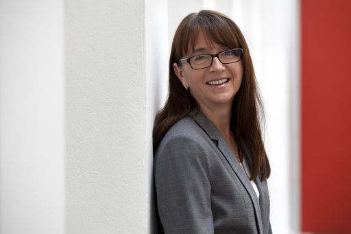 Frau mit braunen Haaren und Brille, grauem Blazer, lächelnd an eine weiße Wand gelehnt.