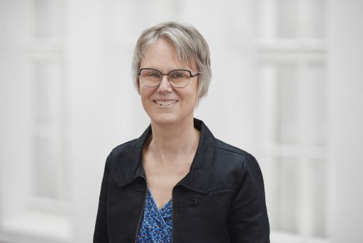 Frau mit kurzen grauen Haaren und Brille, schwarzer Jacke über blauem Muster-Top, lächelt vor hellem Hintergrund.