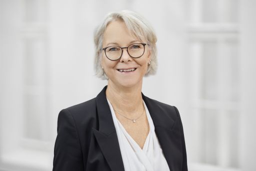 Ältere Frau mit grauem Haar und Brille, lächelnd, in schwarzem Blazer und weißer Bluse, vor hellem Hintergrund.