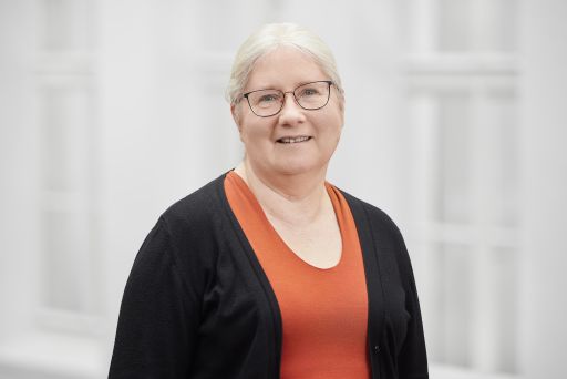 Ältere Frau mit weißen Haaren und Brille, orange Top, schwarzer Strickjacke, lächelt vor hellem Hintergrund.