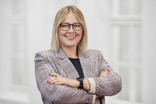 Frau mit blonden Haaren und Brille, im karierten Blazer, Arme verschränkt, lächelt vor hellem Hintergrund.
