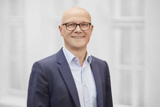 Mann mit Glatze und Brille, blauem Sakko und hellblauem Hemd, lächelt vor hellem Hintergrund.