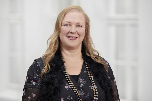 Lächelnde Frau mit langen blonden Haaren, schwarzer Blumenbluse mit Federbesatz und Goldkette vor weißem Hintergrund.