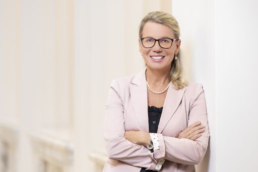 Blonde Frau mit Brille, rosa Blazer und Perlenkette, Arme verschränkt, lächelt, lehnt an heller Wand.