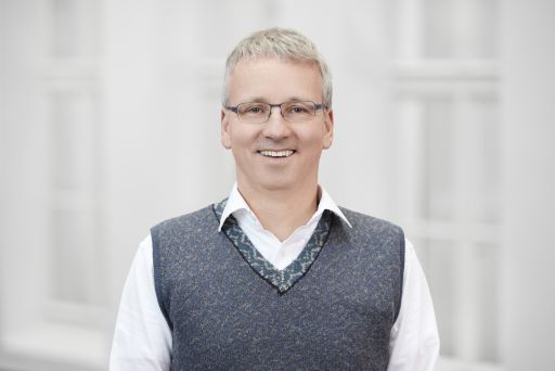 Mann mit grauer Brille und weißen Haaren, trägt weißes Hemd und graue Strickweste, lächelt in die Kamera.