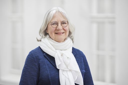 Ältere Frau mit weißem Haar und Brille, blauem Jacket und weißem Schal, lächelt vor hellem Hintergrund.