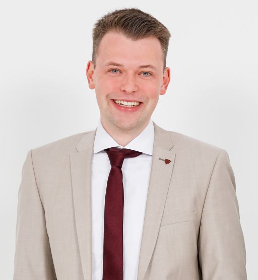 Junger Mann im beigen Anzug mit weinroter Krawatte und Anstecker lächelt vor weißem Hintergrund.