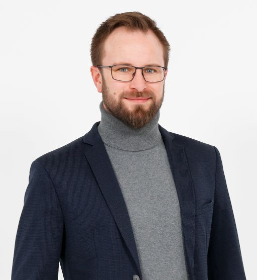 Mann mit Brille und Bart, grauem Rollkragenpullover und dunklem Sakko, vor weißem Hintergrund.