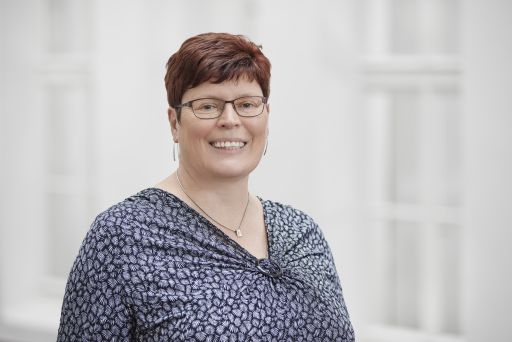Frau mit kurzen roten Haaren und Brille lächelt, trägt gemustertes blaues Oberteil und Halskette.