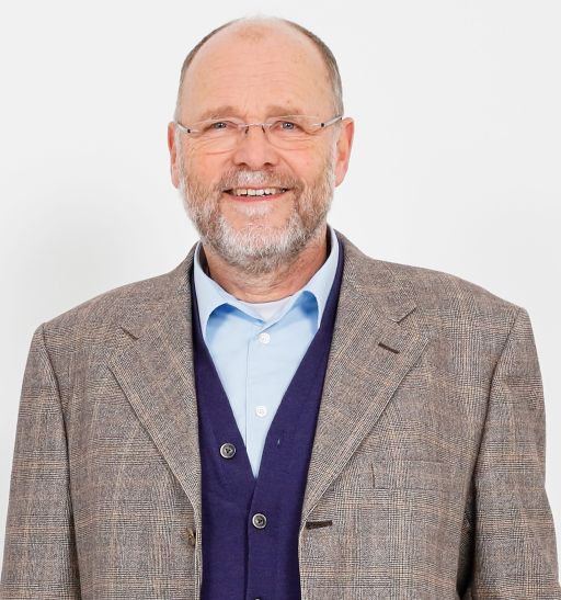 Älterer Mann mit Glatze, Brille und grauem Bart, trägt Sakko, Cardigan und Hemd, lächelt vor weißem Hintergrund.
