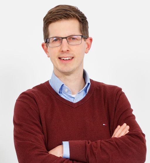 Mann mit Brille, kurzen braunen Haaren, rotem V-Pullover über hellblauem Hemd, Arme verschränkt, weißer Hintergrund.