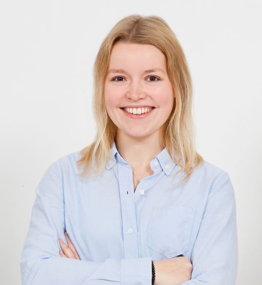 Junge Frau mit blonden Haaren und hellblauem Hemd, Arme verschränkt, lächelt vor weißem Hintergrund.