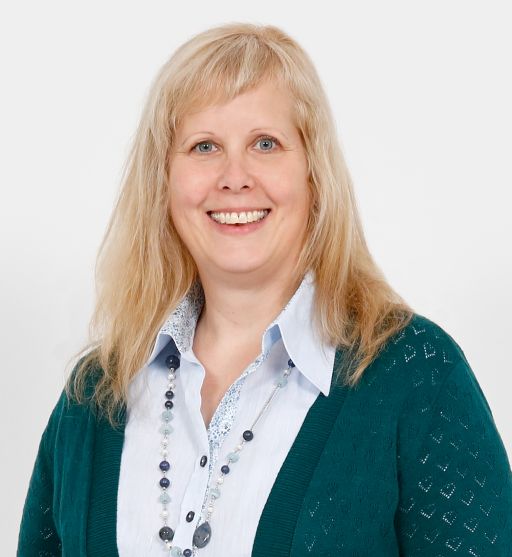 Blonde Frau mit blauen Augen, teal Strickjacke, weißer Bluse und blauer Perlenkette vor weißem Hintergrund.