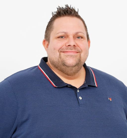 Mann mit kurzem braunem Haar und Bart, lächelnd, trägt dunkelblaues Poloshirt mit roten Streifen, weißer Hintergrund.