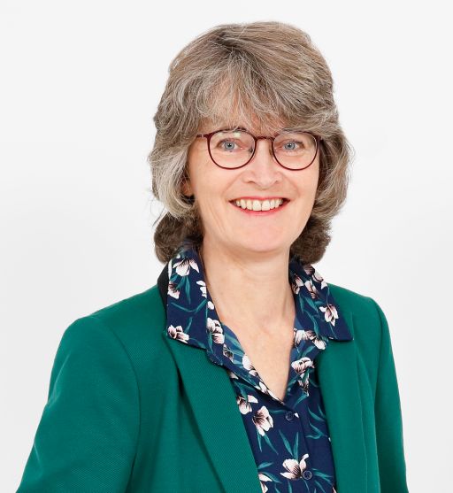 Lächelnde Frau mit grauen Haaren und Brille, grünem Blazer und gemusterter Bluse vor weißem Hintergrund.