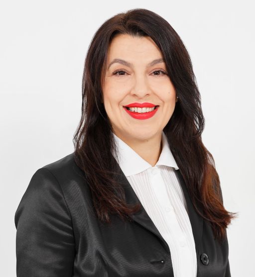Frau mit langen dunklen Haaren, rotem Lippenstift, weißer Bluse und schwarzem Blazer lächelt vor weißem Hintergrund.