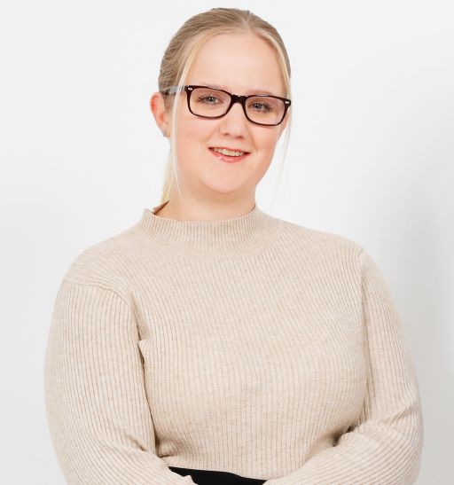 Junge Frau mit blondem Haar, Brille und beigem Strickpullover, Arme verschränkt, lächelt vor weißem Hintergrund.