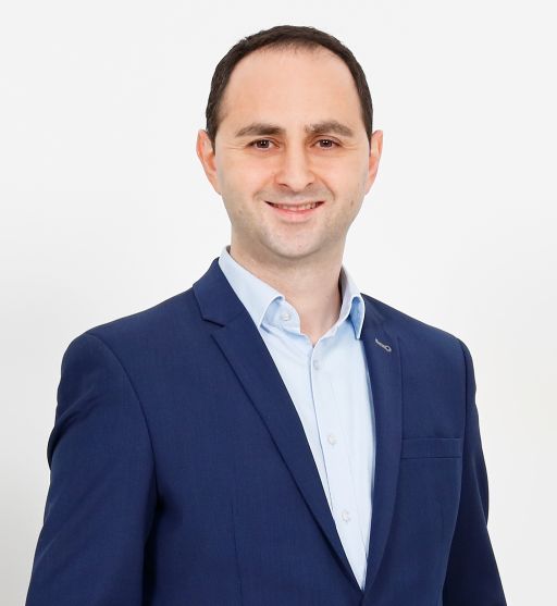 Mann in blauem Sakko und hellblauem Hemd lächelt vor weißem Hintergrund. Professionelles Porträtfoto.