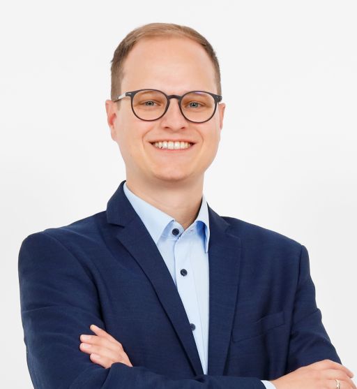 Mann mit Brille und blauem Sakko, Arme verschränkt, lächelt vor weißem Hintergrund.