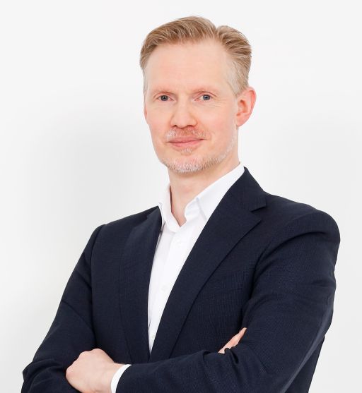 Mann mit blonden Haaren und Bart, dunkles Sakko, weißes Hemd, Arme verschränkt, weißer Hintergrund.