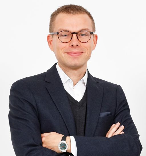 Mann mit Brille und kurzem Haar, Blazer und Uhr, verschränkte Arme, lächelt, weißer Hintergrund.