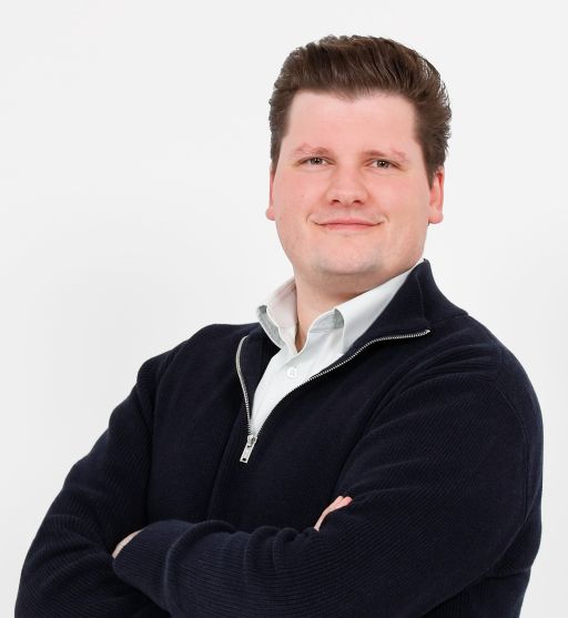 Mann mit dunklem Haar, dunklem Pullover und weißem Hemd, Arme verschränkt, vor weißem Hintergrund.