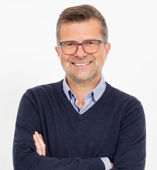 Mann mit Brille, grauem Bart und dunklem V-Pullover über Hemd, Arme verschränkt, lächelt in die Kamera.