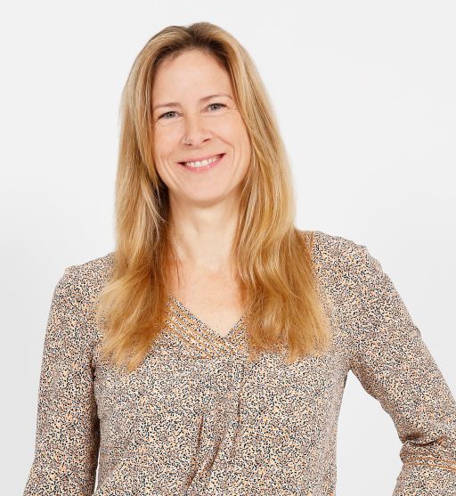 Lächelnde Frau mit langen blonden Haaren in gemusterter Bluse vor weißem Hintergrund.