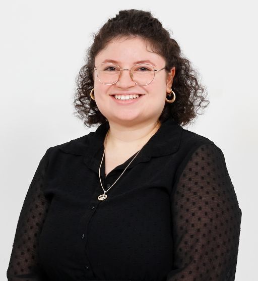 Lächelnde Frau mit lockigem Haar, Brille, goldenen Ohrringen und schwarzer Bluse vor weißem Hintergrund.