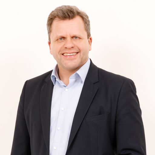 Mann mittleren Alters, kurze braune Haare, lächelnd, dunkles Sakko, hellblaues Hemd, heller Hintergrund.
