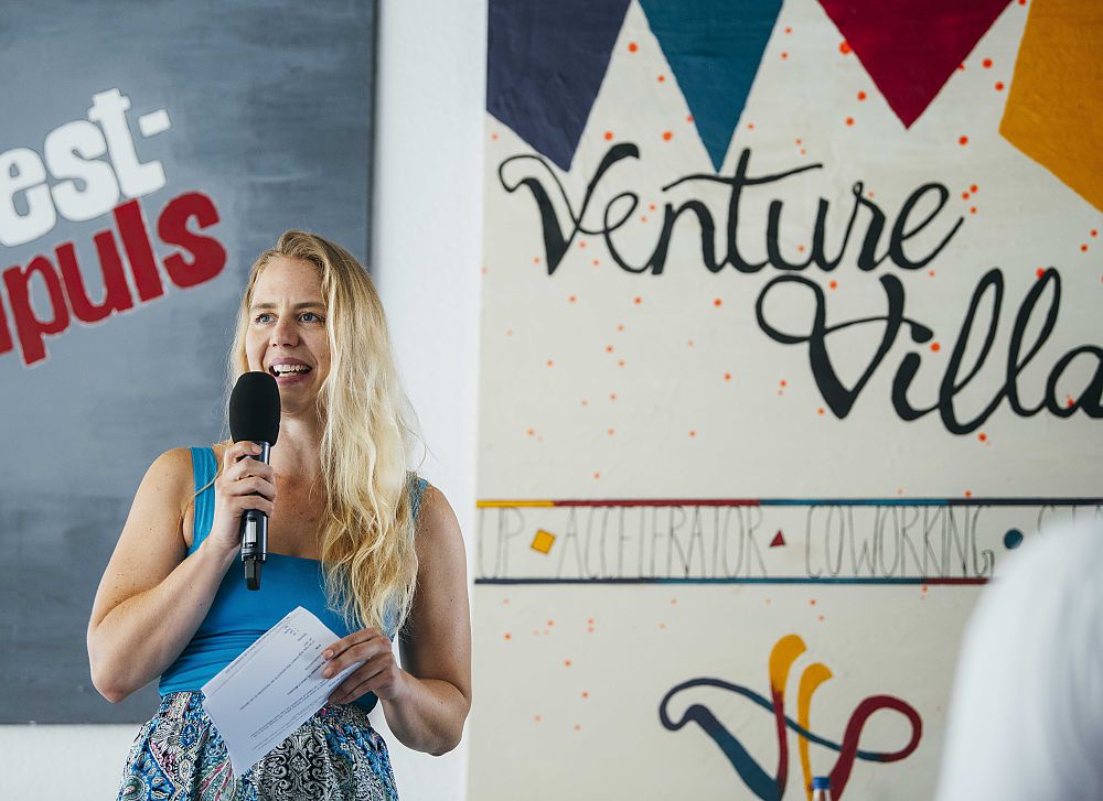 Blonde Frau mit Mikrofon spricht vor einem Venture Villa Schild, hält Papier in der Hand.
