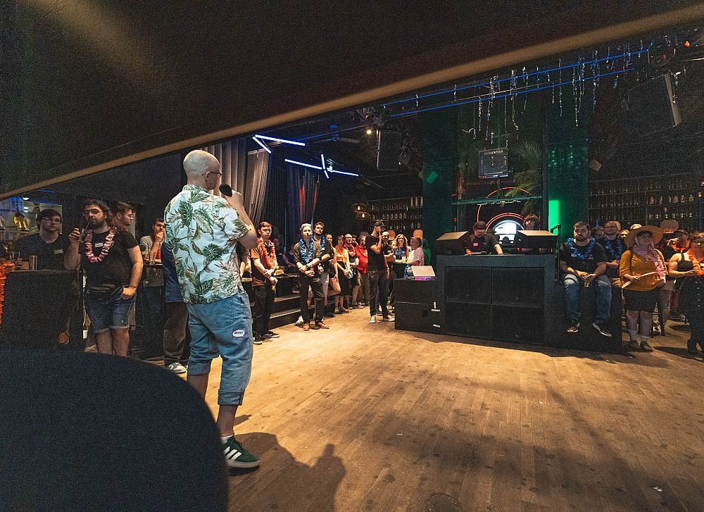 Person in Hawaiihemd steht auf Bühne vor Publikum in dunklem Club mit Holzboden und Neonbeleuchtung.