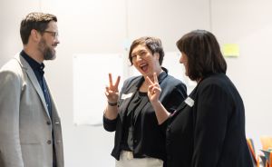 Drei Personen im Gespräch; mittlere Person lacht und zeigt beidseitig Victory-Zeichen. Alle tragen dunkle Businesskleidung.