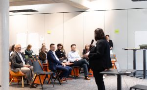 Eine Person hält ein Mikrofon vor einer Gruppe sitzender Zuhörer in einem modernen Konferenzraum.