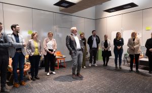 Gruppe von etwa 12 Personen steht im Halbkreis in einem modernen Besprechungsraum mit orangefarbenen Stühlen.