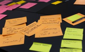 Orangefarbene und pinke Haftnotizen mit deutschen Handschriften auf schwarzem Untergrund, daneben Marker.