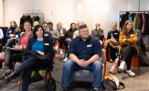 Gruppe von Personen sitzt auf Stühlen in einem Raum und schaut nach vorne, einige tragen Namensschilder.
