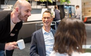 Drei Personen unterhalten sich in einem modernen Büro. Ein Mann hält eine Kaffeetasse, zwei weitere Personen stehen im Hintergrund.