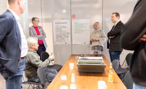 Gruppe von Personen in einem Besprechungsraum mit beschriebenen Whiteboards, Tisch mit Laptop und Getränken.