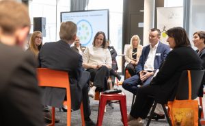 Gruppe von Personen sitzt im Kreis bei einer Podiumsdiskussion in einem modernen Konferenzraum mit Präsentationsscreen.