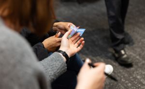 Zwei Personen halten gemeinsam eine kleine blaue Karte mit Text. Eine Person hält einen Stift. Im Hintergrund stehen Beine.