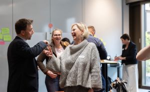 Drei Personen unterhalten sich lachend in einem Büroraum mit Post-its an der Wand; weitere Menschen im Hintergrund.